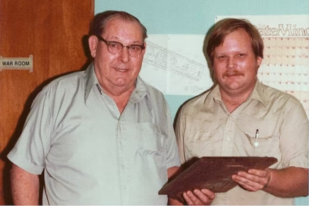 Two men standing indoors in front of a wall with a chart and a door labeled 'WAR ROOM'. The older man on the left wears glasses and a light-colored short-sleeve shirt; the man on the right, with a mustache, holds a wooden plaque.