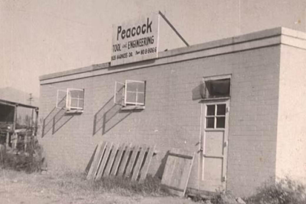 Black-and-white exterior photograph of a single-story brick building with a rooftop sign reading 'Peacock Tool and Engineering.' The building has two windows with metal awnings, a door, and wooden pallets leaning against the wall.
